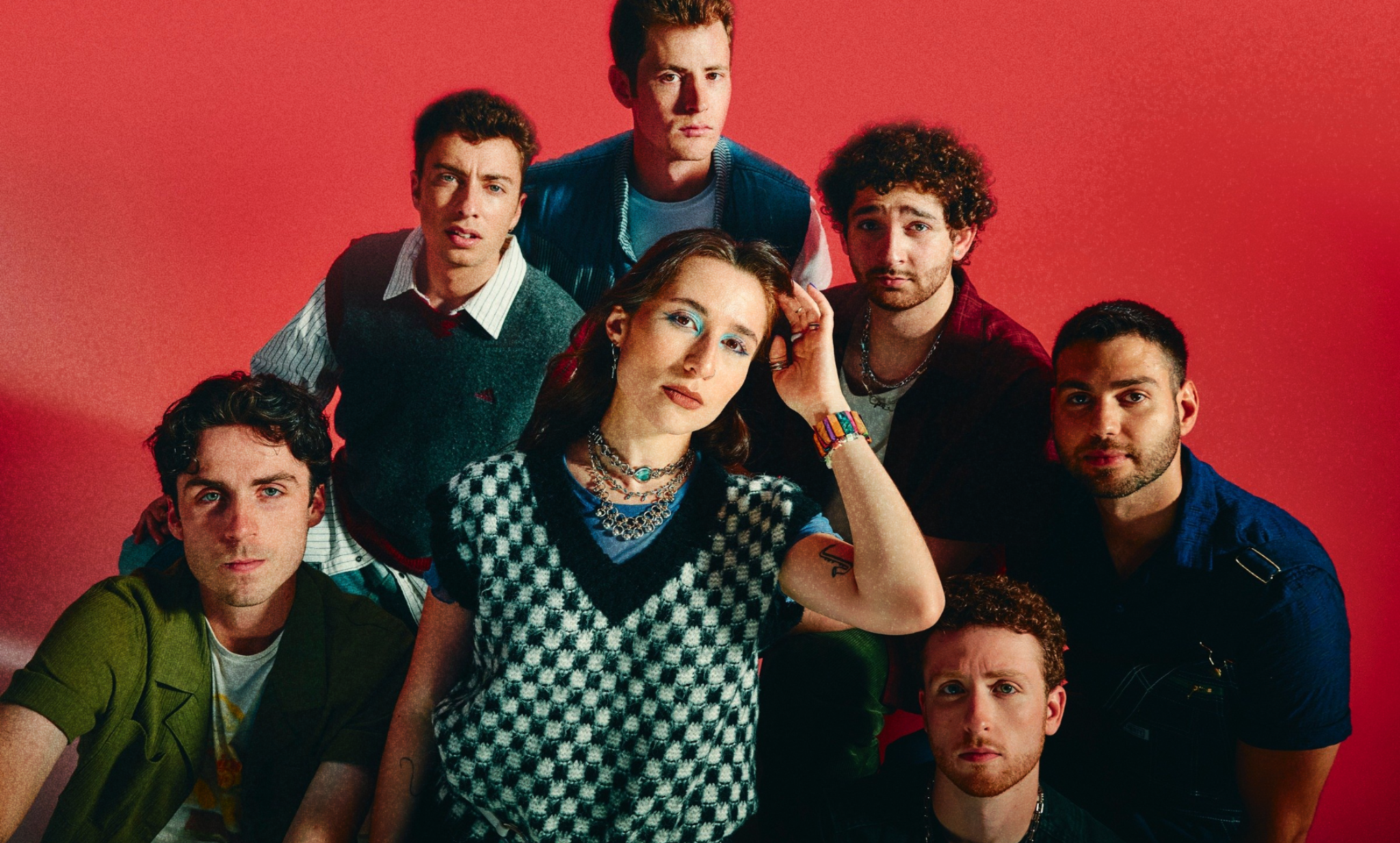 Seven members of the pop band Couch pose together against a red background in colourful retro-inspired outfits
