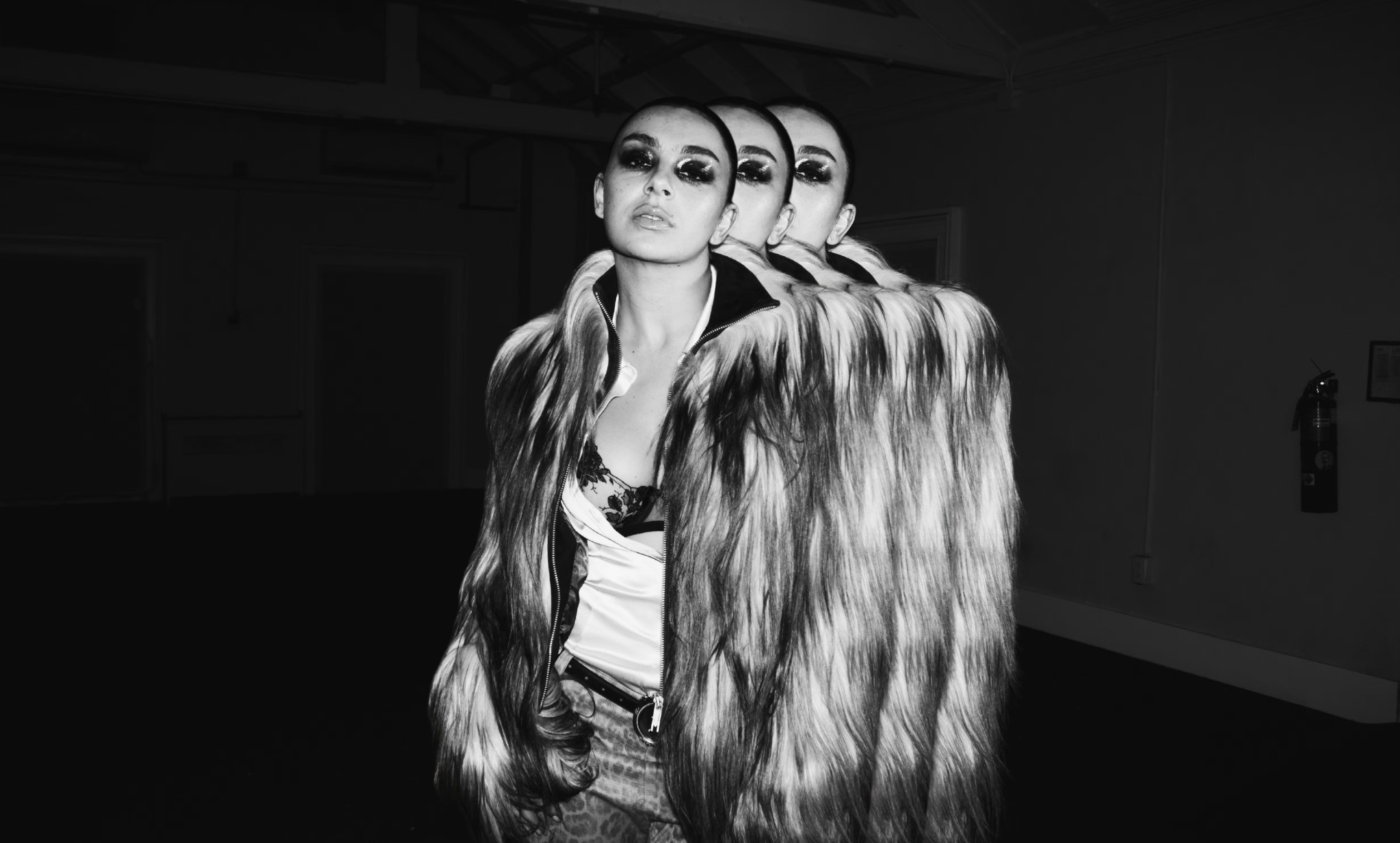 an black and white image of charli xcx by tyrell hampton, with the musician set against a dark backdrop; she's pictured wearing a faux fur coat and dark, bold eye shadow